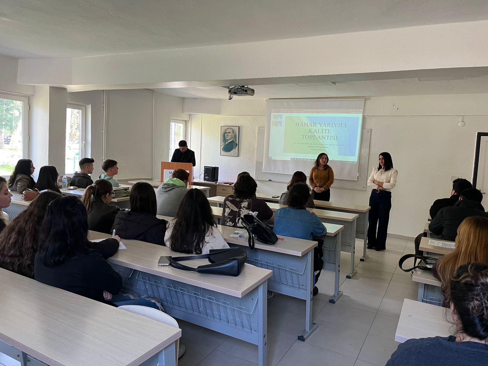 A quality meeting with university students is taking place in a classroom. The instructors are standing at the front of the room and addressing the students. Behind them, a projector screen displays a presentation titled “Spring Semester Quality Meeting.” The students are seated at desks and attentively following the meeting.